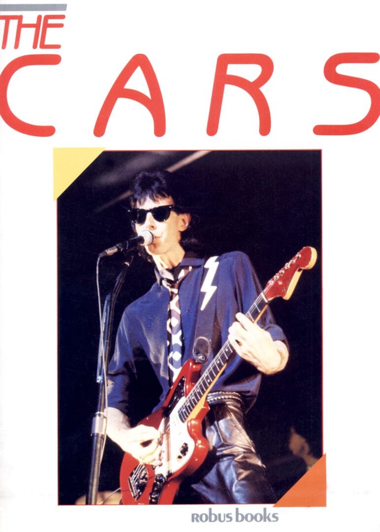 A man holding a guitar on stage with the word cars written above him.